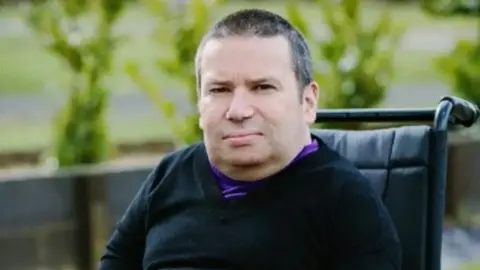 Steve Purse A man wears a black jumper with a purple t-shirt underneath. He is sitting in a wheelchair and looks at the camera.