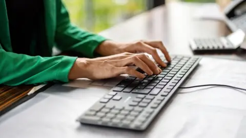 Two hands resting on a black keyboard. The keyboard is on a dark wooden desk. 