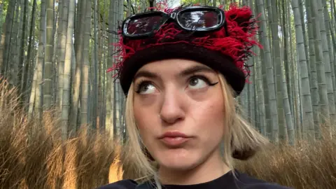 Izabel Rose Izzy, wearing a black and red fluffy hat and glasses perched on her head, with giant bamboo in the background 