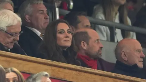 The Prince and Princess of Wales in the stands at the Principality stadium watching Wales v England Six Nations match. Kate is on the left and looking out to the field smiling. Williams is on the right and is looking away from the camera. 