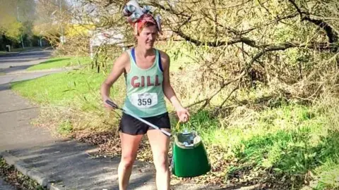 Gill Thomas Gill Thomas picking litter while out on a half marathon. She is wearing running gear - a vest with her name on it and shorts. She is carrying a bag and a litter pick.