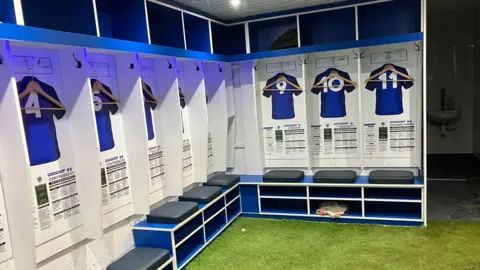 Inside a football dressing room. Small areas for number 4 to 11 are listed. Each area has a padded seat, storage, and a coat hanger for the match shirt. The ground is covered in artificial turf, and there are blue lights illuminating the room