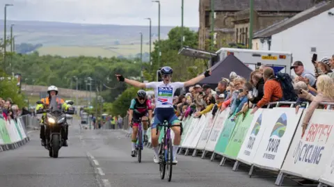 A female cyclist throws her arms in the air as she crosses the finishing line of the race on 22 June. Another cyclist and a man on a motorbike are following behind. Spectators are lining both sides of the street.
