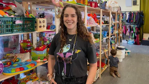 Hannah Jones stands in front of a shelf of toys. She has brown curly hair and is wearing a black tee-shirt featuring Disney character Maleficent. 