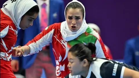 Three women are involved in the game. Two women are wearing red and white kits and have headscarves on, a third is captured lower down in the photo and is wearing a black and white kit. 