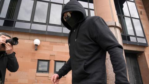 A man wearing a black face covering and jacket with his hood pulled over his head, pictured outside court