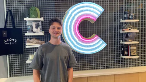 A man with short black hair is wearing a grey t-shirt. He is stood by a wall which has small black tiles. There is a large letter C in blue and pink neon lights and items including shoes and bags on small shelves