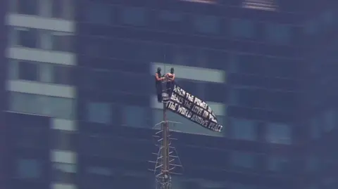 Two small figuress in black at the very top of the spire, with windows a building behind them, a black flag with white script waves to one side
