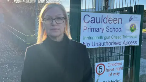 Jackie Hendry has blonde hair and is wearing glasses. She is wearing a black coat over a black top. She is standing at the gates of Cauldeen Primary School.