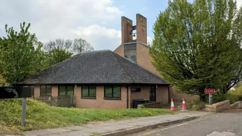 Fabian Musto / geograph A small brick church with a low, dark roof and a tall bell tower stands beside a quiet street, surrounded by green trees and grass.