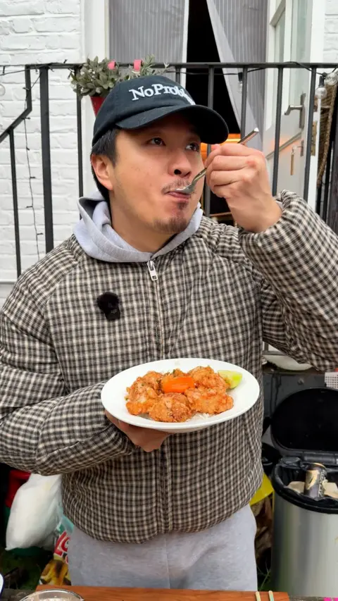 A man in a checked top and baseball cap eats from a spoon while holding a bowl of food