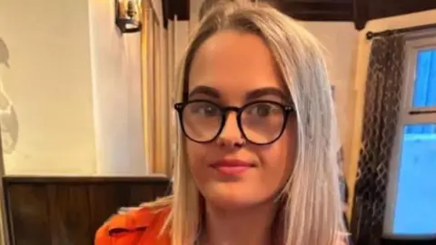 Tegan Williams Tegan, with bobbed bleach blonde hair and dark glasses. Her shoulders are just visible, she wears an orange jacket. 