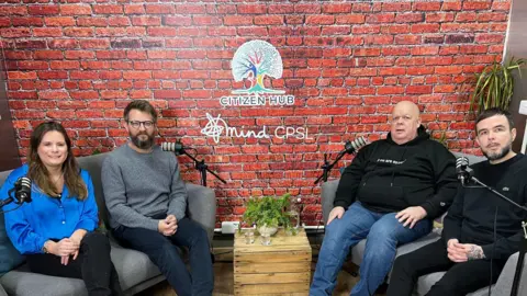 CPSL Mind Members of the CPSL podcast team - a woman in a blue blouse, a man in a grey jumper and navy trousers, a man in a black hoodie and blue jeans and a man in a black jumper and black trousers - sitting on grey sofas on either side of the room with microphones in front of them.