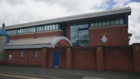 An outside view of Bolton Police Station, which is a multi-storey brick and glass building with blue window frames, and surrounded by a brick wall. A sign reading Bolton Police Station and a GMP crest are mounted to the side of the building. 