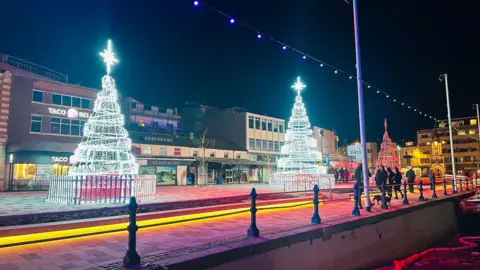 Three large metal Christmas trees sit on the harbourside. They have metal fencing around their bases and giant stars on the top of them. People are walking by and it is night-time. There are shops and restaurants in the background including Taco Bell and Debenhams. In the foreground are iron bollards and there are fairy lights running along the harbourside.