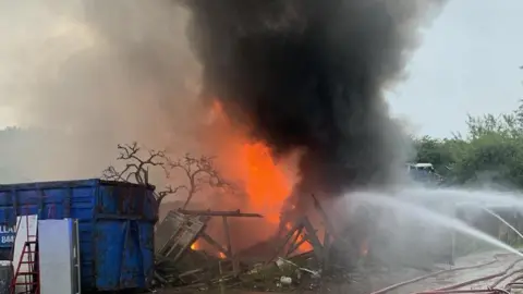 Water hoses directed towards an area a few metres wide of orange flames and smoke next to a blue skip