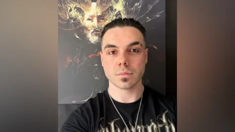Damien Powell Damien Powell has dark slicked back hair. He is wearing a band tee-shirt and standing in front of some gold artwork depicting a face. He has a nose and lip piercing and a chain around his neck. 