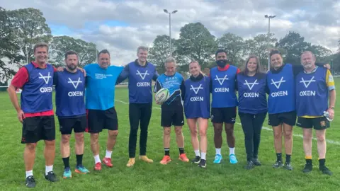 BBC Gary Shaughnessy centre is smiling at the camera, wearing black shorts and a blue t-shirt whilst holding a rugby ball.