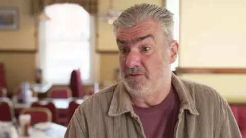 A man with grey hair, slicked back with hair product, wears a beige cord shirt and burgundy t-shirt underneath. He is being interviewed in his fish and chip restaurant, with red and gold chairs in the background.