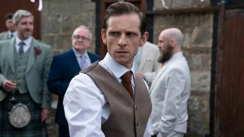 Actor Jamie Bell in a scene in the TV series Half Man. He is wearing a white shirt, a red tie and a brown waistcoat, in what seems to be a wedding scene - other men are dressed in suits and kilts in the background in front of a brick building. Bell has a deeply concerned look on his face.
