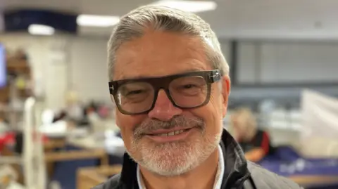 The managing director or FaberExposize UK stands in the printing factory that he runs. He smiles at the camera and is wearing glasses.