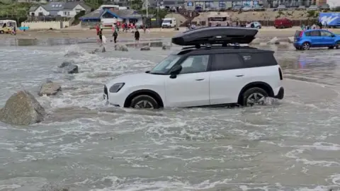A white Mini with a black roof box is surrounded by the sea with the water half- way up its wheels. There is no-one in the car. There are large boulders placed in front of it as markers. In the background are a couple of parked cars and vans parked on the wet beach. There are also people walking along the beach towards the buildings facing onto the beach.