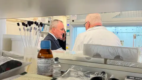 Conor Murphy speaking to another man with his back to the camera wearing a white lab coat. Conor Murphy is wearing a suit and tie. In the foreground a shelf with laboratory equipment on it