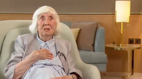 Betty Brown wearing a blue dress, grey jacket and pearls sits in a green armchair. In the background is a lamp on a table.
