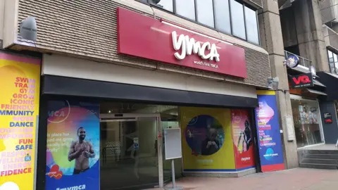 An exterior view of the YMCA building. Above the entrance is a large red sign saying YMCA World's first YMCA and the windows either side to the entrance have colourful blue and posters on them.