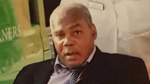 an older man, dressed in a black jacket, lilac shirt and striped tie is sat facing the camera