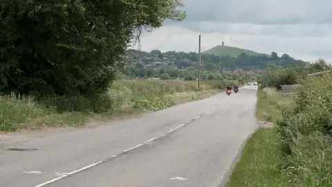 A straight country road with motorcyclists driving along and cars further away. Beyond the trees in the far distance is Glastonbury Tor.