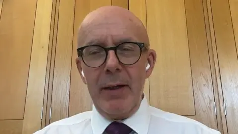 A bald man with black glasses and white shirt and claret tie sat in an oak panelled room