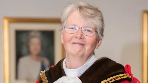 City of Wolverhampton Council Claire Darke in a portrait. She is wearing glasses and smiling. She has her mayoral dress on and there is a blurred picture of the queen behind her. 