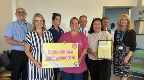 CDDFT A group of eight hospital staff wearing a mixture of healthcare and office outfits smile at the camera in a hospital setting. They are holding a yellow sign that reads 'Sophie's Legacy Approved Hospital'