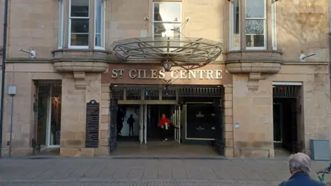 The front of the St Giles Centre in Elgin. A woman in a red coat walks through the open doors. 