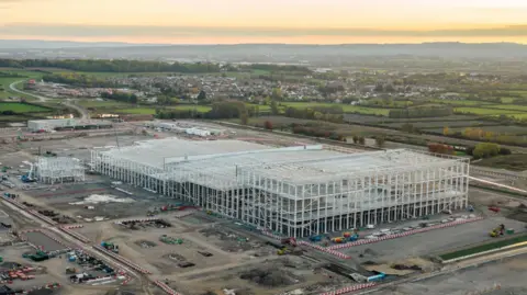 An aerial photo at sunset of the huge gigafactory steel frame on its construction site with the Somerset countryside surrounding it