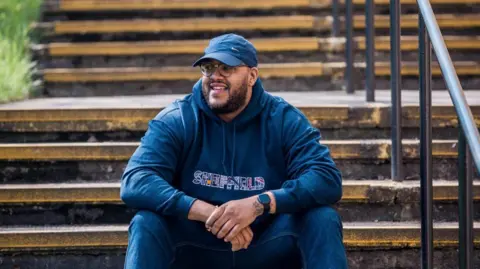 Image shows a man with a beard and wearing glasses and a blue baseball top sitting on some stairs outdoors