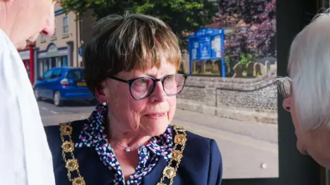 North Norfolk District Council Victoria Holliday is wearing a gold ceremonial chain over a navy blue jacket. Beneath that, she has a patterned blouse. She is talking to a man and a woman.