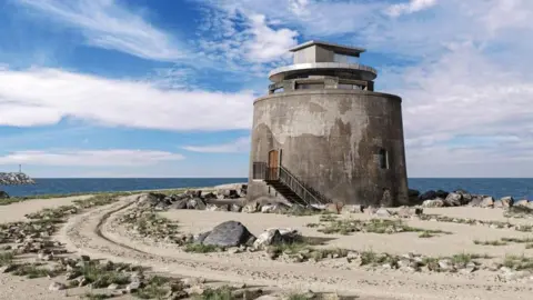 A CGI picture of a large cylindrical stone structure on a beach with the sea in the background