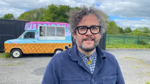 Elizabeth Baines/BBC A man in a blue painter's jacket has grey hair. Behind him an ice cream van is parked up.