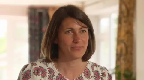Kate, a woman with short brown hair and wearing a pink floral dress. She is smiling. The background is out of focus but a green house plant and some curtains can be seen. There are also white window frames and it looks to be a bright day outside.