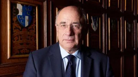 Alamy A head-and-shoulders portrait of Sir Brian Leveson, a white man with rimless glasses and grey hair at the sides of his head, wearing a navy suit with a light blue shirt and navy tie. He is standing in a wood-panelled room with heraldic designs behind him and is light by soft light coming from the left of the picture.