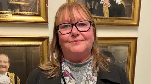 A woman with long hair, glasses and wearing a black coat with a scarf and grey top. Stands in front of a wall with four framed portraits.