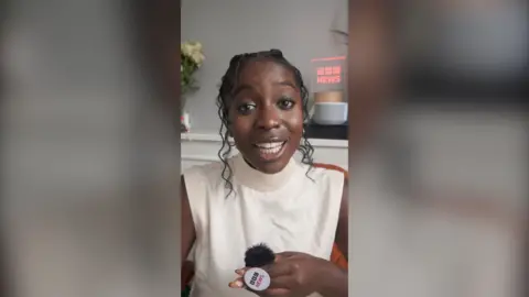 Reporter Jonelle Awomoyi is wearing a white sleeveless turtleneck. She has curly black hair. She is holding a BBC button mic.