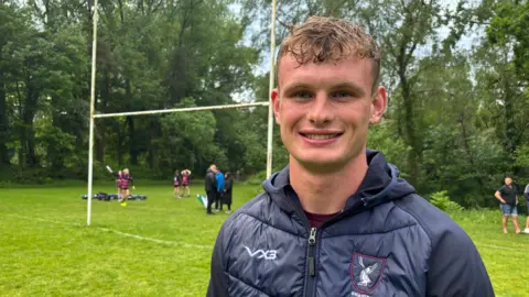 Joshua smiles at the camera. He has short, curly hair is wearing a navy down jacket with a club badge on the right hand side. There is a rugby pitch behind him where there are children and adults standing by the rugby goals.