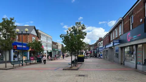A pedestrianised High Street with trees, benches and paving. There are shops on both sides of the street. It is a sunny day with blue sky.