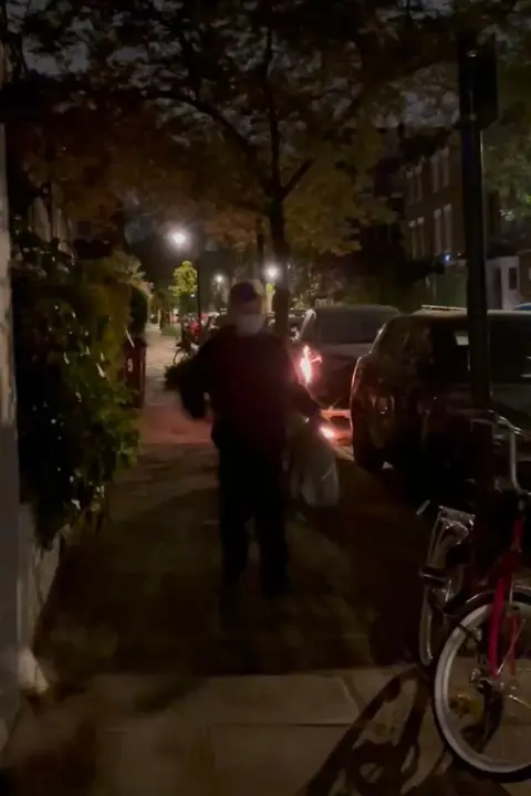 Counter Terrorism Police A figure dressed in dark clothing and a white face mask stands on a pavement in the dark, next to him is a bicycle and a car, behind him another car appears to be on fire.