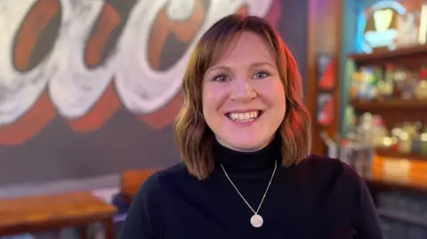 Charlie Kemp is smiling with a blurred out background behind her. She has shoulder length light brown hair and is wearing a silver pendant and dark blue polo neck jumper.