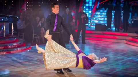 Una foto de Widdecombe y Du Beke bailando en el estudio de Strictly Come Dancing. Anton Du Beke lleva camisa y pantalón negros y corbata morada. Hace girar a Ann Widdecombe, sujetándola de la mano y del pie. Widdecombe lleva un vestido dorado brillante con mangas moradas.