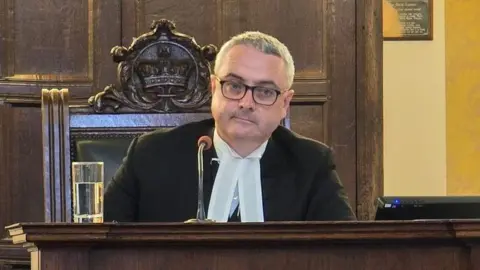Watterson in a black jacket and white shirt sitting at the Leg Co a wooden bench in the chamber.